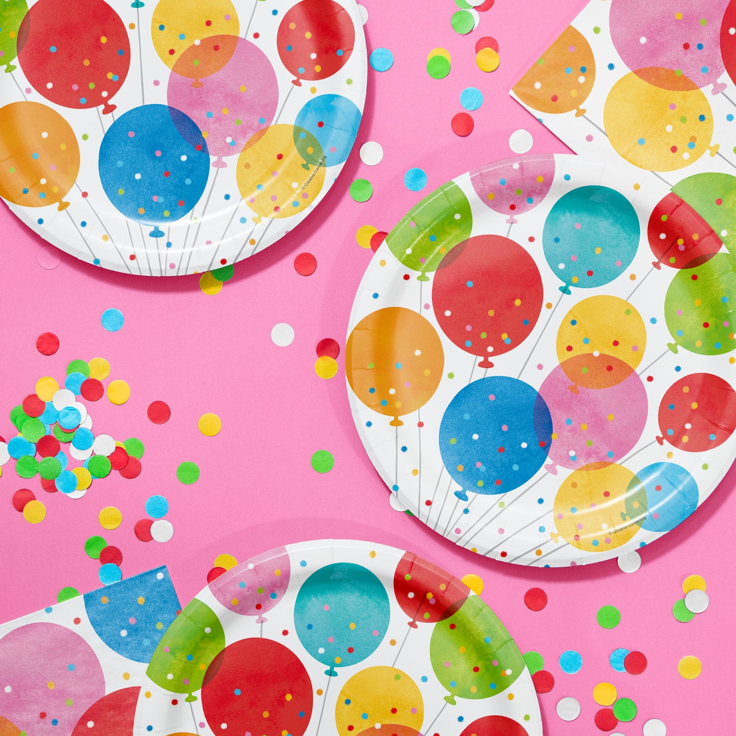 Colorful paper plates with balloon design on a pink background with colorful confetti.