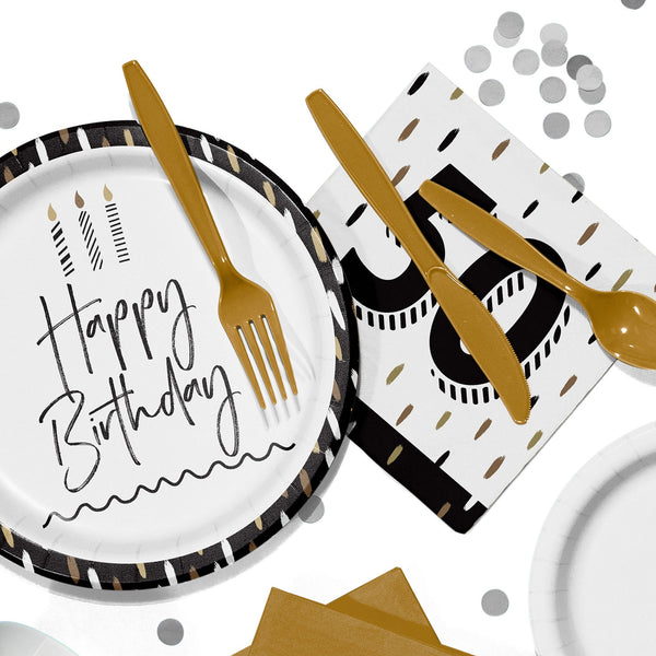 Flat lay of black, white, and gold birthday party tableware, including a paper plate that reads “Happy Birthday,” gold plastic cutlery, a napkin with a large “50,” and scattered silver confetti on a white background.