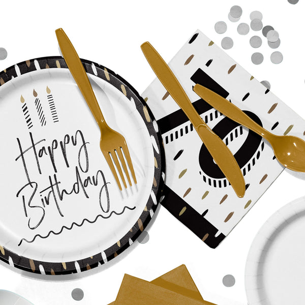 Flat lay of black, white, and gold birthday party tableware, including a paper plate that reads “Happy Birthday,” gold plastic cutlery, a napkin with a large “70,” and scattered silver confetti on a white background.