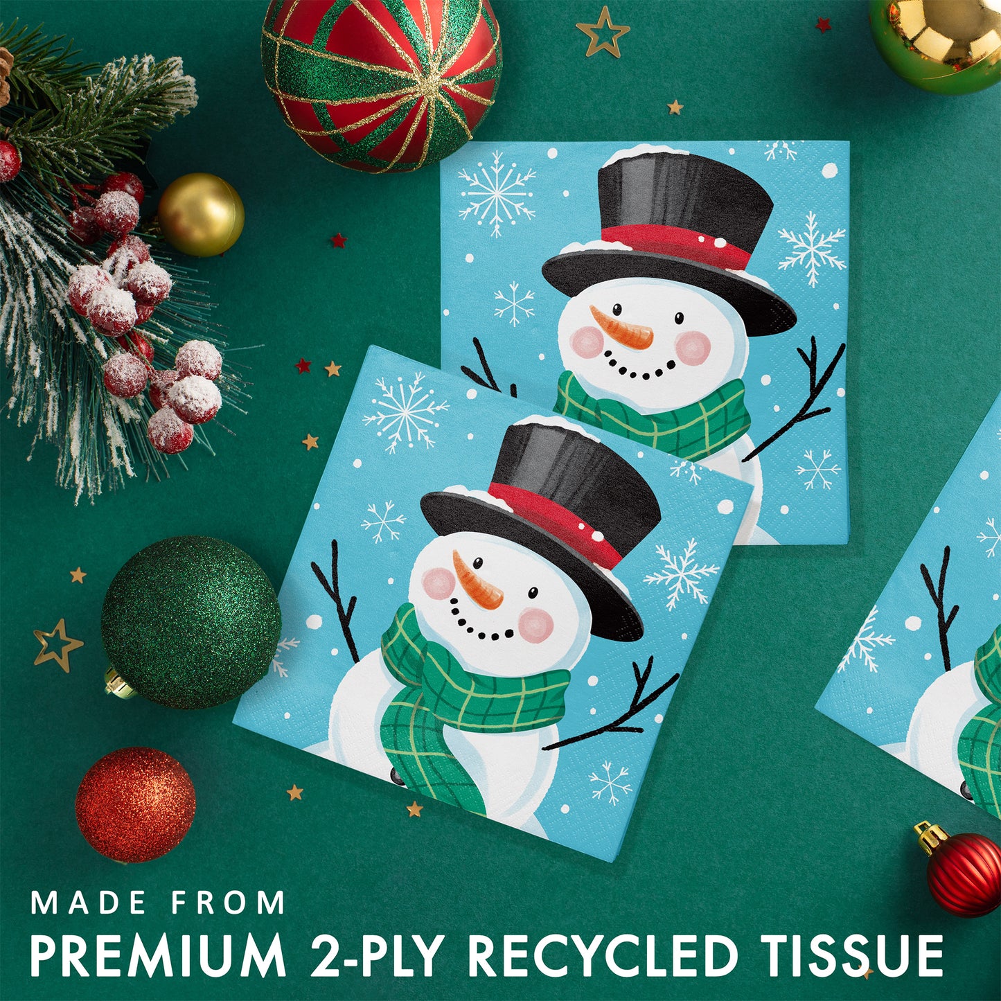 Two Christmas-themed paper napkins with a snowman and snowflakes on a blue background placed on a green background and Christmas decorations.