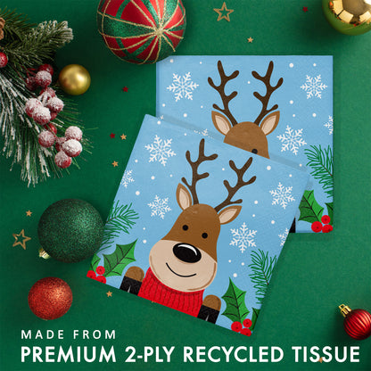 Christmas Cheer Luncheon Napkins 192 ct. Paper napkins featuring a cartoon reindeer with a red sweater on a blue background with snowflakes and greenery. Shown on a green background with ornaments and greenery.