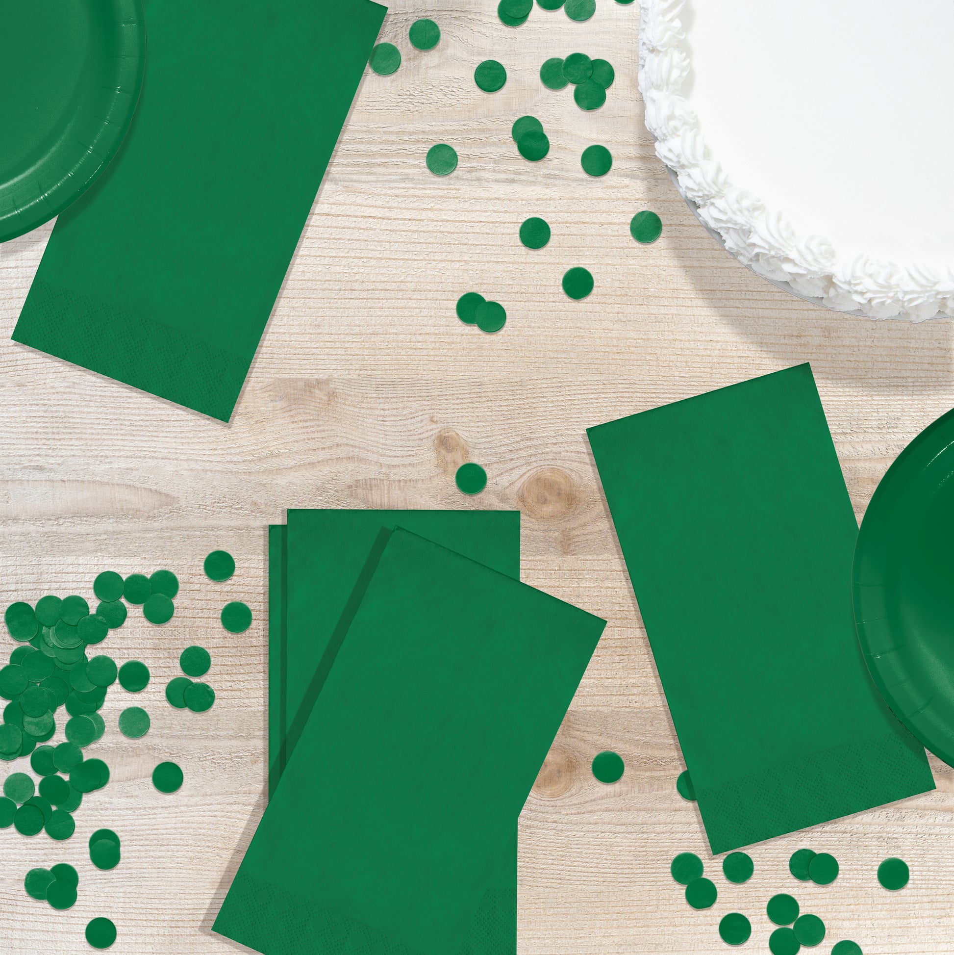 Green party supplies including napkins and plates on a wooden table with scattered green confetti.
