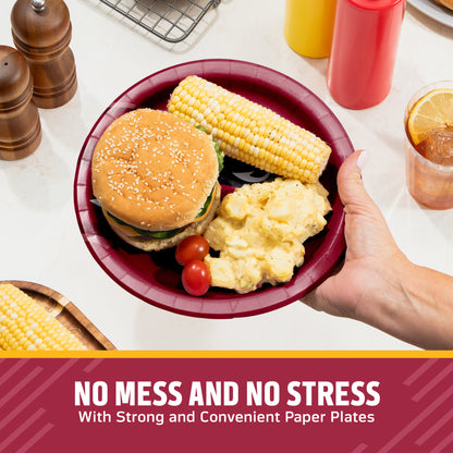 A hand holds a dark red paper plate with a hamburger, corn on the cob, mashed potatoes, and cherry tomatoes on a kitchen counter. Condiment bottles, pepper mills, and a drink are visible in the background, with a text banner at the bottom reading 'NO MESS AND NO STRESS With Strong and Convenient Paper Plates'.