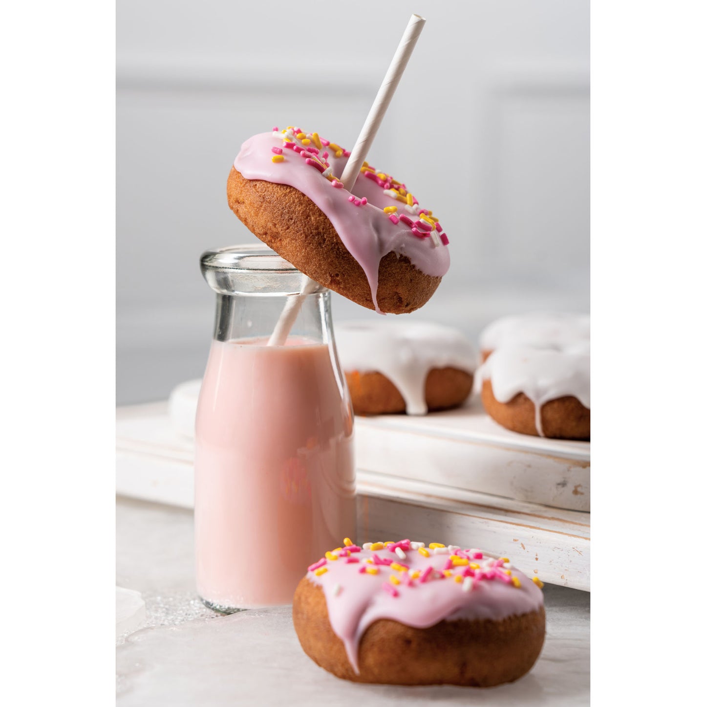 Donuts with pink glaze and sprinkles next to a bottle of pink milk with a white paper straw on a light wooden surface.
