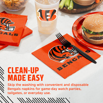 Cincinnati Bengals disposable napkins and matching drink cup set on a table, with a burger, plate, fork, and cleanup promotional text visible. The napkins are bright orange with the Bengals helmet logo and team name printed on them.