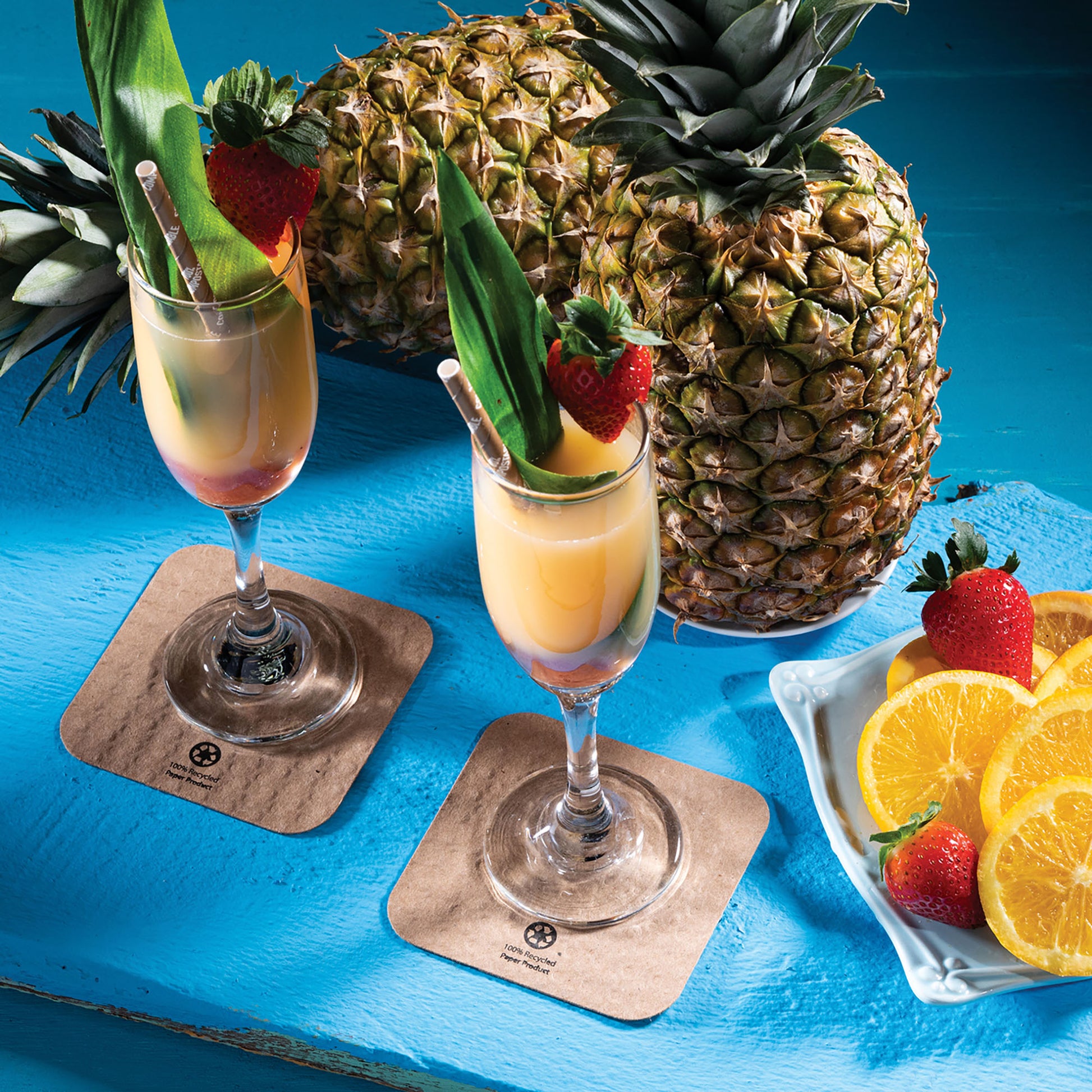 Fruity drinks shown on coasters surrounded by pineapples, oranges and strawberries on a blue tablecloth