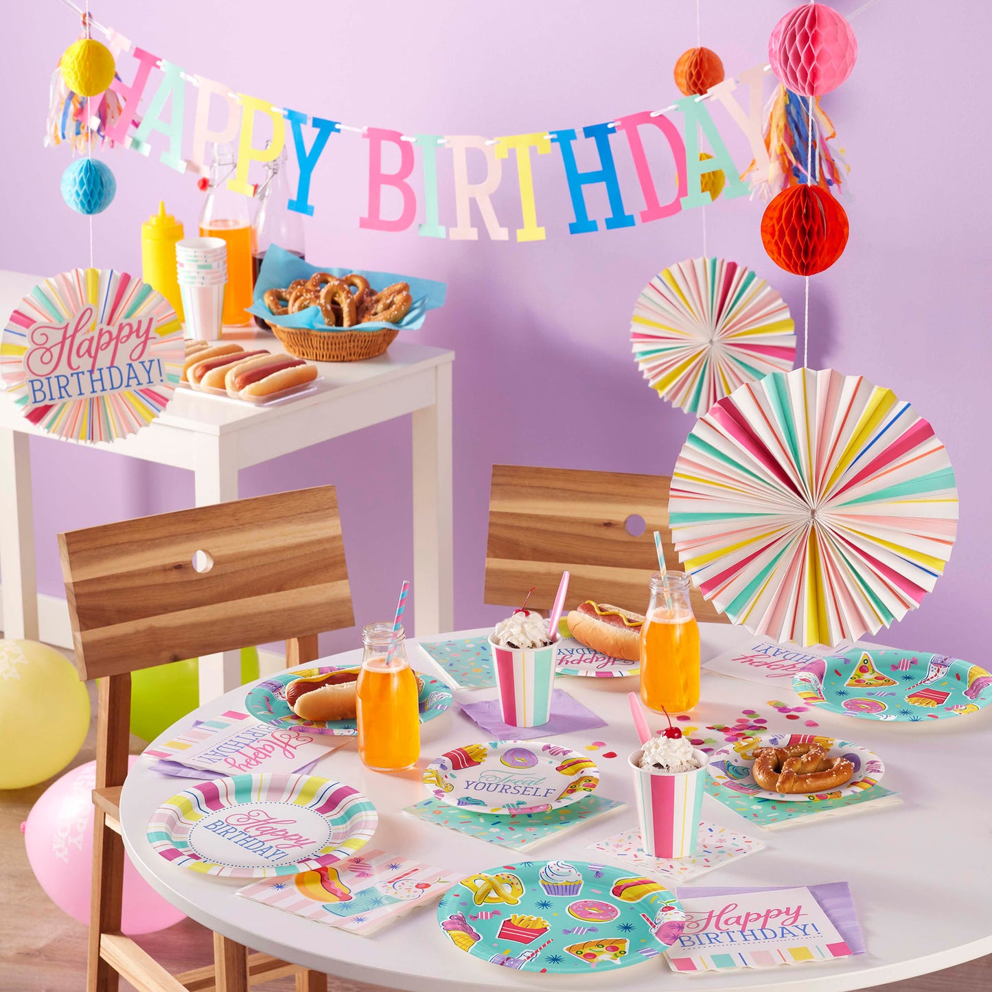 Birthday party setup with colorful decorations, tableware, and food in front of a purple wall