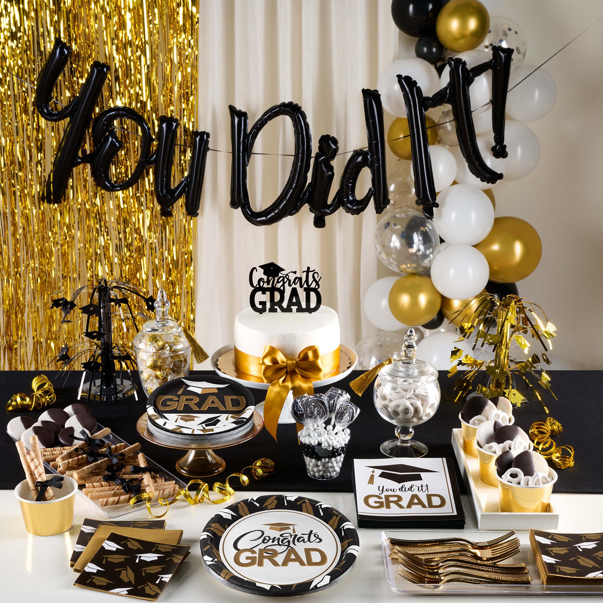 Graduation-themed table setup with balloons, cake, and decorative items against a gold and black backdrop.