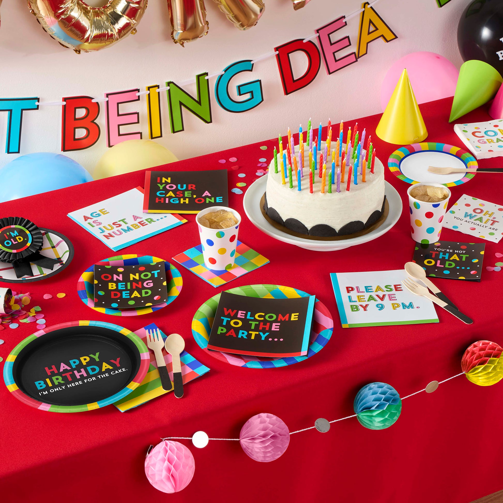 Birthday party setup with cake, plates, and decorations on a red tablecloth.